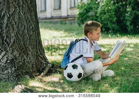 Cute Smiling Schoolboy Reading Book While Sitting On Lawn Near Soccer Ball