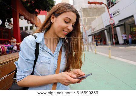 Beautiful Student Girl Sitting On Street Bench Messaging With Mobile Phone In Sao Paulo City, Brazil
