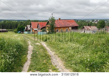Village Street In Summer, Overgrown With Grass And Flowers. View Of The Traditional Wooden Rural Hou