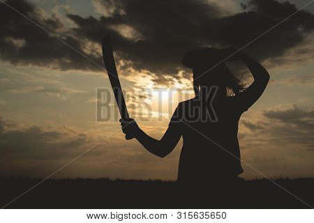 Silhouette Of Pirate Woman In A Pirate Hat And With A Sword In Hands Over A Evening Cloudy Sky Backg