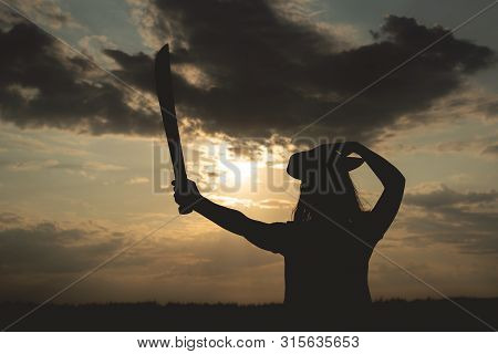 Silhouette Of Pirate Woman In A Pirate Hat And With A Sword In Hands Over A Evening Cloudy Sky Backg