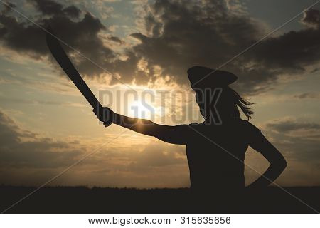 Silhouette Of Pirate Woman In A Pirate Hat And With A Sword In Hands Over A Evening Cloudy Sky Backg