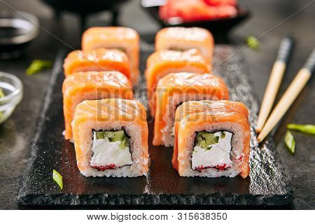 Beautiful Philadelphia Sushi Rolls Set with Salmon and Cream Cheese, Tobiko and Cuccumber on Black Slate Plate Close Up. Uramaki, Nori Maki or Futomaki Sushi with Trout Fillets, Soy Sauce and Wasabi