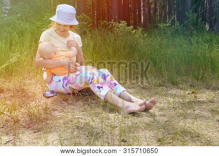 Smiling Mother Sits On The Shore Of A Forest Lake On A Sunny Summer Day, Holds Her Little Son In Her