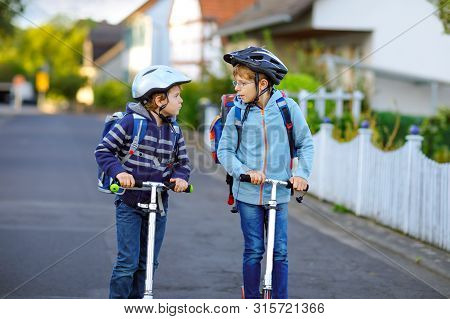 Two School Kid Boys In Safety Helmet Riding With Scooter In The City With Backpack On Sunny Day. Hap