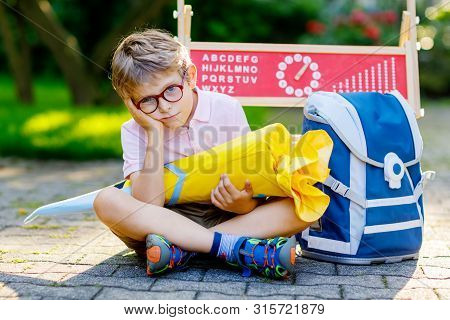 Frustrated Sad Little Kid Boy With Glasses Sitting By Desk And Backpack Or Satchel. Schoolkid With T