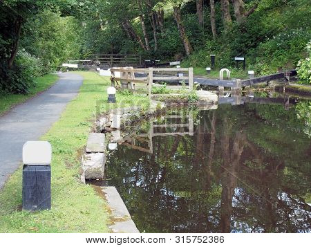 A Footpath Neext To Old Wooden Lock Gates With Trees And Flowers Reflected In The Water With Mooring