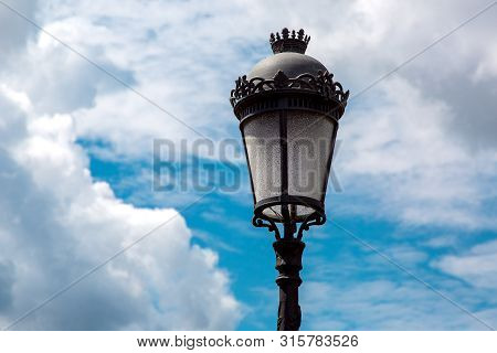 Black Iron Street Lamp With A Pattern And A Glass Shade Against The Sky With Clouds On A Sunny Day.