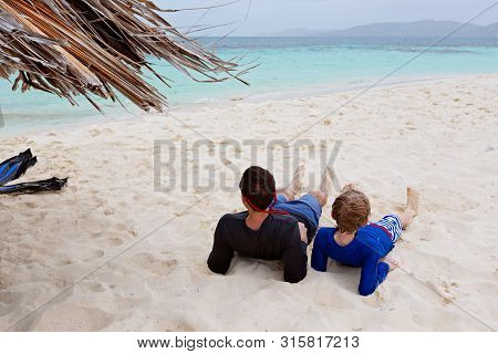 Family Of Two, Father And Son, In Sunglasses And Rashguards Lying At The Beach Enjoying Beautiful Tu