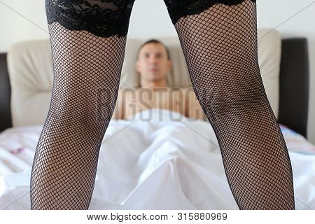 Couple Of Lovers In A Bed, Foreplay Before Sex. Girl In Black Fishnet Stockings Standing In Front Of