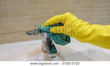 Woman In Yellow Rubber Gloves Cleaning A Water Tap. Housewife Holding A Rag. White Sink. Modern Bath