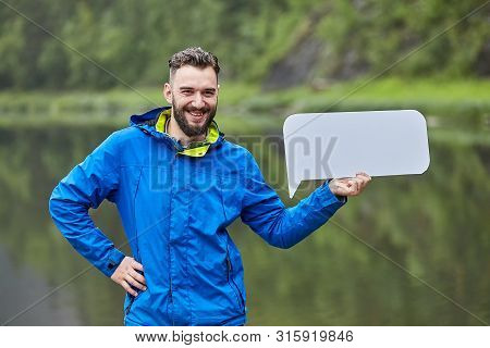 Cheerful Young Caucasian Man Is Holding Blank Banner Ont The Backdrop Of A Natural Landscape.
