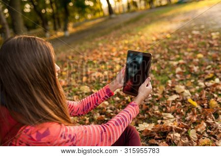 Pretty young girl using tablet outdoor lying on leaves and smiling. Woman using digital tablet pc in the autumn park. Student using tablet after school.