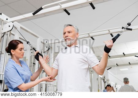 Brunette Rehabilitologist Assisting Middle Aged Man Working Out In Hospital Gym.