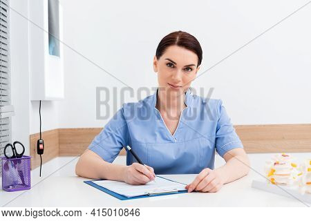Rehabilitologist Looking At Camera While Writing Prescription On Clipboard.