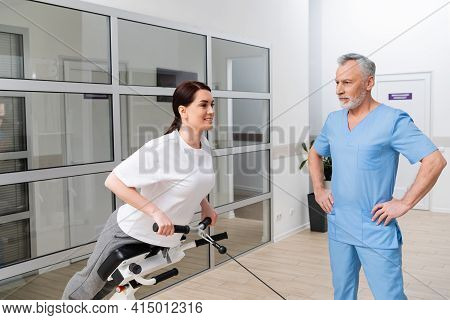 Middle Aged Rehabilitologist Standing With Hands On Hips Near Smiling Woman Training On Exercising M