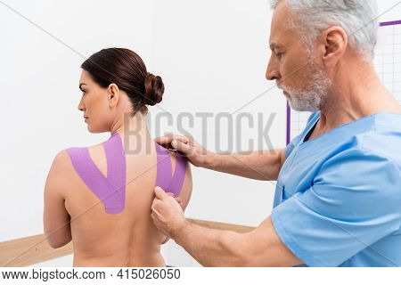 Mature Rehabilitologist Applying Tape On Back Of Woman During Kinesiotherapy.
