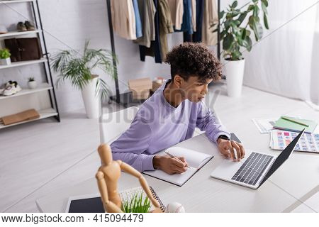 Young African American Retailor Writing On Notebook And Using Laptop At Table.