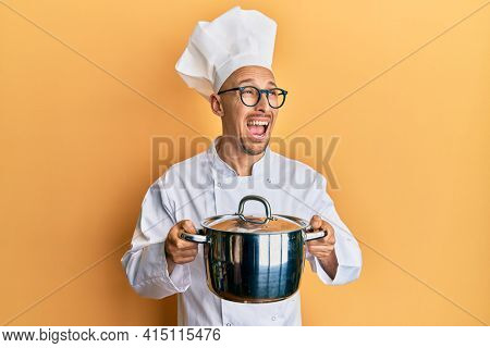 Bald man with beard wearing professional cook holding cooking pot angry and mad screaming frustrated and furious, shouting with anger. rage and aggressive concept. 