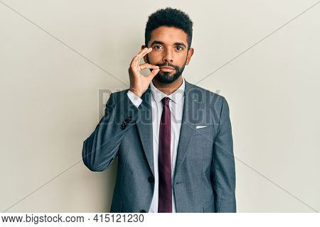 Handsome hispanic man with beard wearing business suit and tie mouth and lips shut as zip with fingers. secret and silent, taboo talking 