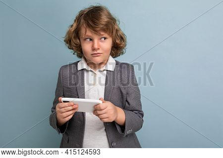 Photo Shot Of Handsome Suspacted Child Boy With Curly Hair Wearing Grey Suit Holding And Using Phone