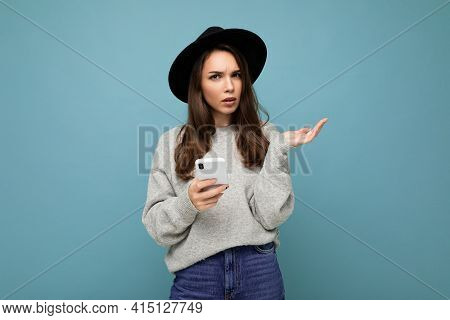 Beautiful Young Brunette Woman Thinking Wearing Black Hat And Grey Sweater Holding Smartphone Lookin