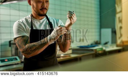 Young Chef Standing In The Cuisine In Gloves. Asian Cook In Modern Cook Room. Bearded Male In Apron.