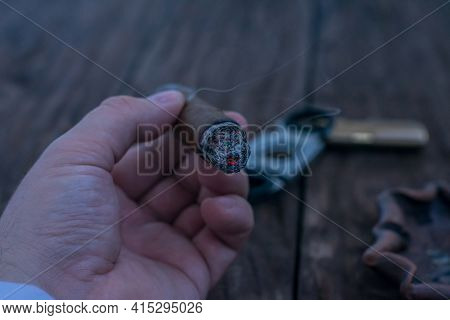 A Man's Hand Holds A Smoldering Cigar Over A Wooden Table Against The Background Of A Guillotine, A 
