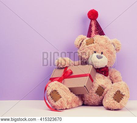 Cute Brown Teddy Bear In A Red Cap Sits And Holds A Brown Box With A Gift, Festive Background, Copy 