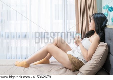 Asian Glasses Woman Holds A Cup Of Coffee And Relax On The Bed In The Morning At Her Bedroom.