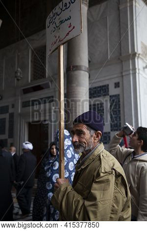 Damascus, Syria 03/28/2010: An Old Bearded Man Wearing A Rain Coat And A Winter Cap Is Holding A Dir