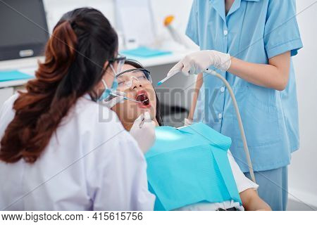 Nurse Putting Saliva Ejector In Mouth Of Female Patient When Dentist With Mirror And Tweezers Checki