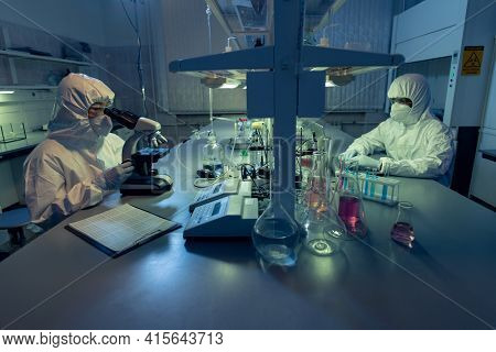 Two colleagues in protective workwear sitting at the table and working with chemical samples in test tubes in the laoratory