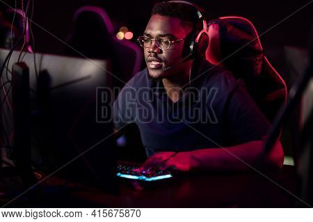 An African Student Sits In A Gaming Chair In A Computer Club, Puts On Headphones, Smiles, And Prepar