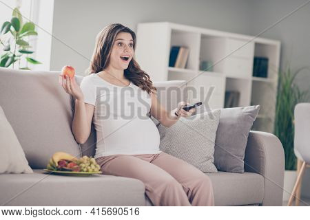 Photo Of Attractive Young Lady Hold Fruit Sit Sofa Watch Tv Motherhood Amazed Film Indoors Inside Ho
