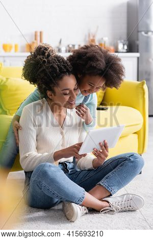 Smiling African American Woman Sitting On Floor And Using Digital Tablet Near Excited Child.