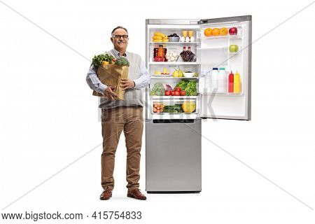 Full length portrait of a mature man with a grocery bag standing next to an open full fridge isolated on white background