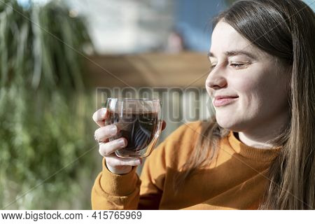 Smiling Young Pretty Caucasian Woman In Orange Sweatshirt Holding Glass Cup Of Coffee Or Cocoa In Su