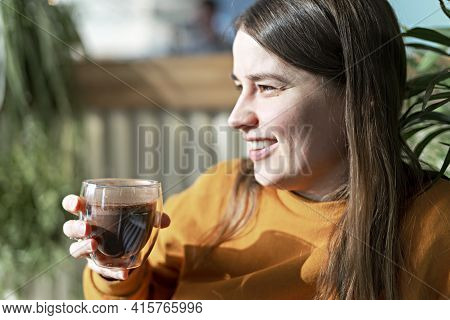 Smiling Young Pretty Caucasian Woman In Orange Sweatshirt Holding Glass Cup Of Coffee Or Cocoa In Su