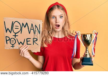 Beautiful young caucasian girl holding woman power banner and trophy afraid and shocked with surprise and amazed expression, fear and excited face. 