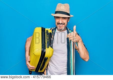 Middle age handsome tourist man on vacation holding cabin bag over isolated blue background smiling happy and positive, thumb up doing excellent and approval sign