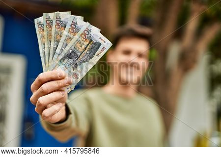 Young hispanic man smiling happy holding peruvian soles banknotes at the city.