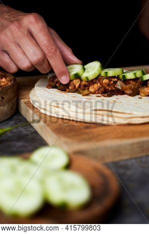 a young caucasian man is filling a durum or a burrito, with chicken meat cooked with different vegetables such as onion or red and green pepper, and some slices of raw cucumber