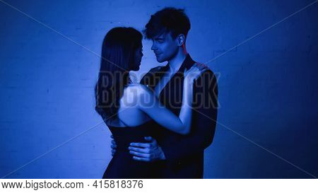 Seductive Woman In Black Dress Embracing Man In Blazer On Blue.