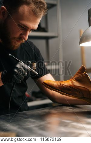 Portrait Of Bearded Shoemaker Wearing Black Gloves Spraying Paint Of Light Brown Leather Shoes, Clos