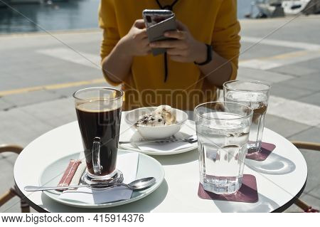 Mug Of Coffee, Water Glasses And Bowl Of Ice Cream On Seaside Cafe Table With Woman Sitting By And U