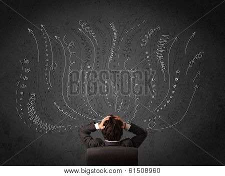 Thoughtful young businessman sitting and deciding in front of a chalkboard with sketched arrows and lines in different directions