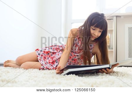Charming little girl reading book on white carpet and smiling