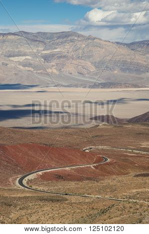 Winding road going down into Panamint Valley, passing through a red cinder cone, with cloud shadows on the valley floor.