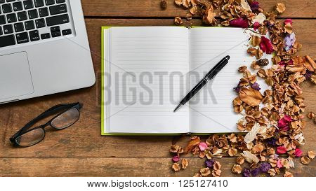 Top view workspace with notebook, pen, glasses, laptop and dried flowers on wooden table background.